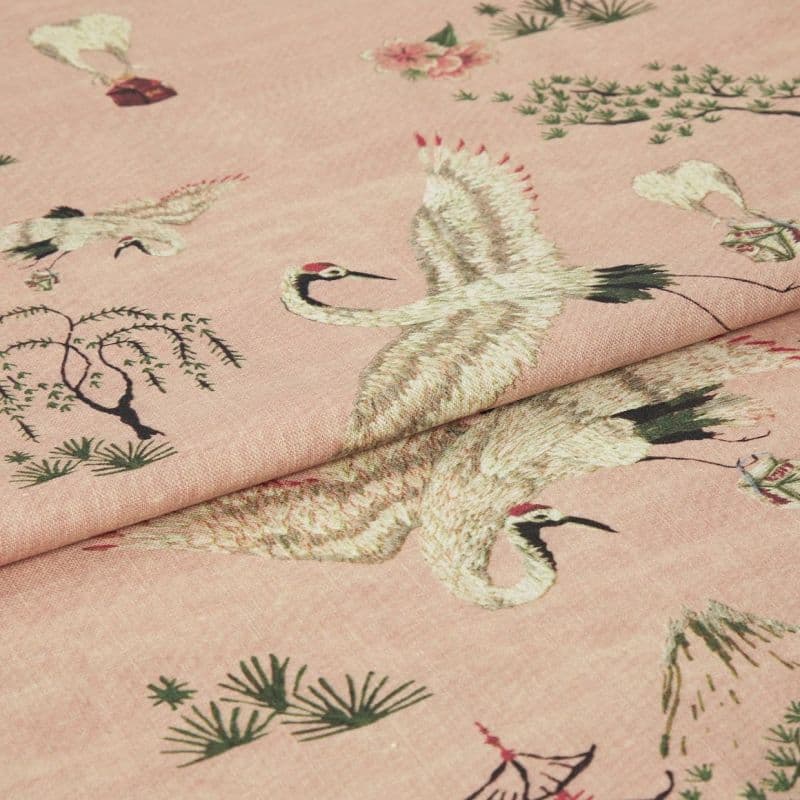 Pink woven fabric printed with red-crowned cranes and stylized trees — folded to show layered panels — close-up on a soft textile surface under soft, even lighting.