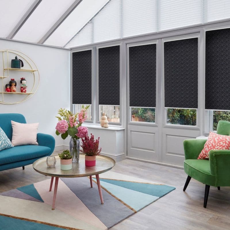 Black patterned roller blinds on tall conservatory windows and French doors, lowered to cover upper panes, filtering light; bright sunroom with glass roof, colourful sofas, rug, coffee table and plants.