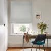 Light-gray Roman blind covering the window, lowered almost fully with a small gap; filtering soft daylight. In a neutral home office: wooden desk with mirror, vase of flowers, teal chair.