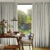 Full-length pleated pale-gray curtains with subtle floral sprigs, parted at center to reveal the glass door and filter soft daylight; dining table set for breakfast in a sunlit garden-facing room.