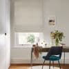 pale textured Roman blind lowered mid-window, folded at bottom, filtering soft daylight; desk with mirror, vase of flowers and teal-upholstered chair in a neutral study.
