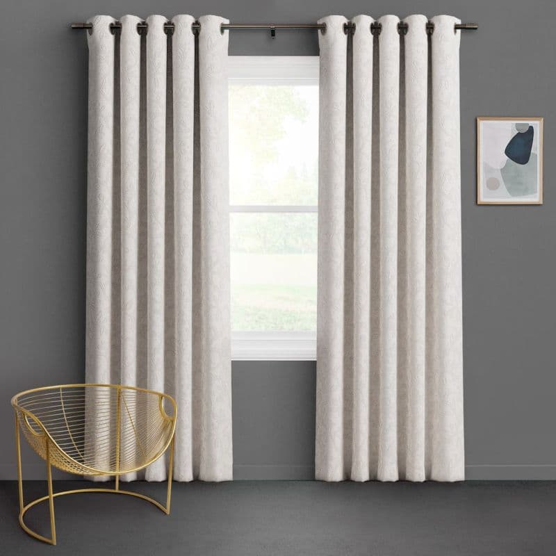 Cream patterned grommet-top curtains, hung on a metal rod and slightly parted to reveal a bright window, filtering daylight in a gray-walled room with a gold wire chair.