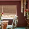 Woven beige patterned roman blind lowered over a window, filtering daylight with its bottom slightly raised; in a burgundy dining room with brass-framed floral chairs, wooden table, and beaded pendant lights.