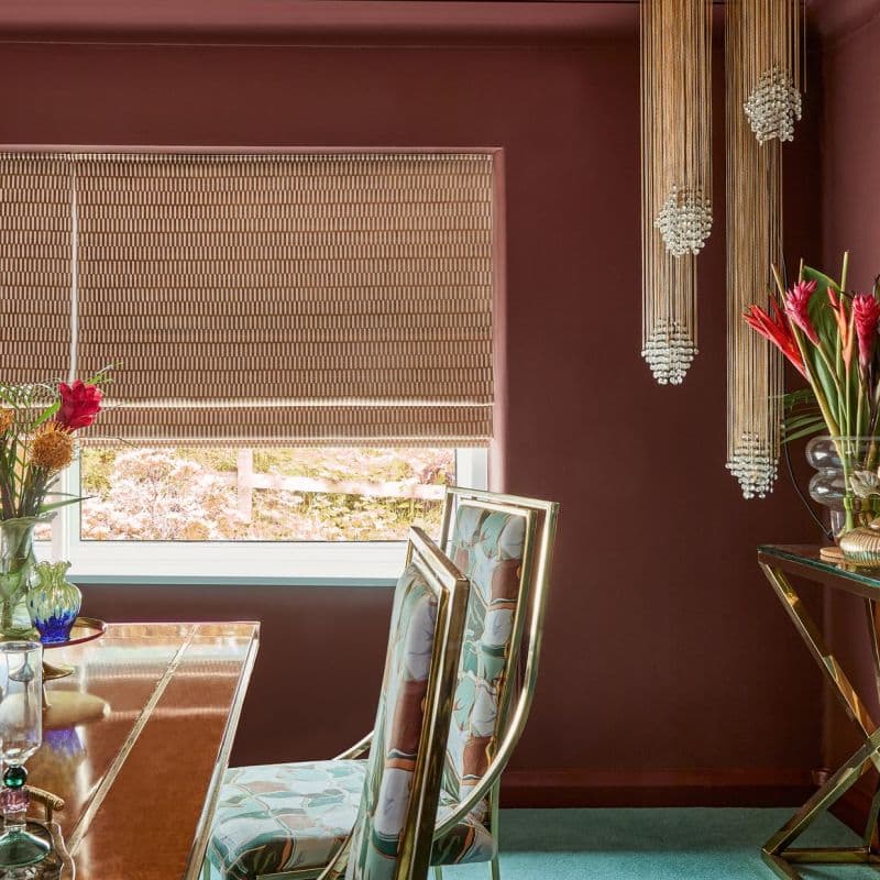 Woven beige patterned roman blind lowered over a window, filtering daylight with its bottom slightly raised; in a burgundy dining room with brass-framed floral chairs, wooden table, and beaded pendant lights.