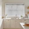 White horizontal Venetian blinds across a large window, slats mostly closed and filtering soft daylight into a bright, neutral-toned kitchen with sink, marble countertop, and small plants.
