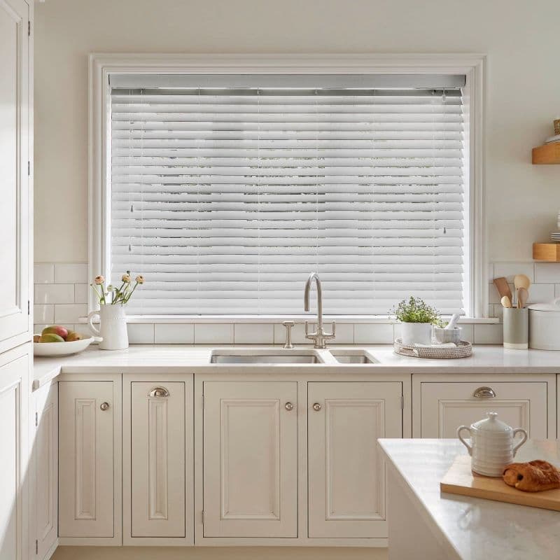 White horizontal Venetian blinds across a large window, slats mostly closed and filtering soft daylight into a bright, neutral-toned kitchen with sink, marble countertop, and small plants.