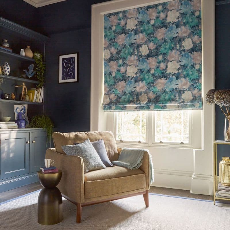 Floral blue-green Roman shade, fully lowered with watercolor-like blooms, filtering soft daylight above a white-framed window in a dark-blue-walled living room reading nook with an upholstered armchair and shelves.