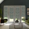 Patterned floral roller blinds cover large glass French doors, partially lowered and filtering light, in a living room with green armchairs, glass coffee table, chevron rug, and plants.