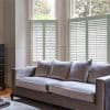 White plantation-style café shutters covering the lower halves of tall windows, slatted panels partially closed and filtering daylight; grey sofa with cushions in a lit living room, trees visible outside.