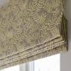 Roman blind with beige fabric and yellow fern-like pattern, neatly folded into pleats; lowered over a window, partially blocking and softly filtering daylight in a neutral-toned room.