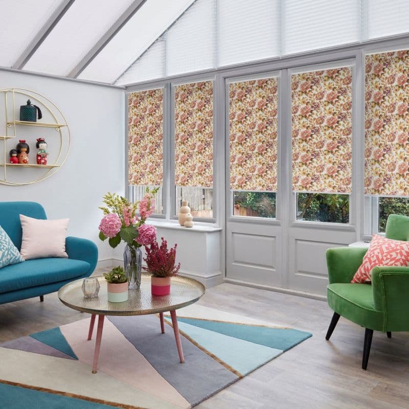 Floral-patterned roller blinds lowered over tall conservatory windows and doors, filtering daylight; modern sunroom with teal sofa, green armchair, geometric rug and coffee table topped with bouquets.
