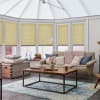 Honey-yellow pleated cellular blinds, fitted across bay conservatory windows and lowered mid-height, filtering soft daylight; modern cozy sitting area beyond with beige sofa, chunky pink throw, wooden coffee table.