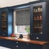 Roman fabric blind with floral pattern, lowered to cover most of the window and filter light; set within navy kitchen cabinetry above a wooden countertop with vase and fruit bowl.