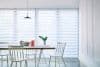 Zebra blinds cover floor-to-ceiling windows, alternating translucent and opaque stripes lowered to softly filter daylight, in a bright minimalist dining room with a wooden table, white chairs and a plant.