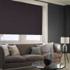 Dark fabric roller blind lowered nearly to the sill, blocking light over a two-pane living-room window; charcoal walls, beige sofa with cushions, glass coffee table and side lamp.