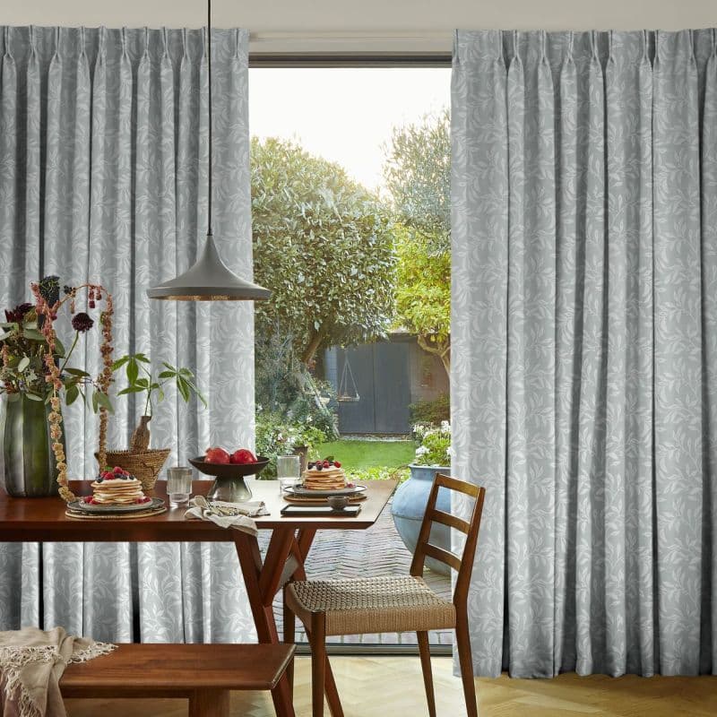 Light-grey, floor-length pleated curtains with subtle leaf pattern, parted at center; letting soft daylight through; framing a dining table set for breakfast and a sunlit backyard with trimmed trees.