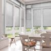 Grey horizontal Venetian blinds cover tall glass doors and windows, slats mostly closed and filtering daylight; bright white dining area with wooden table, chairs, floral cushions and garden view.