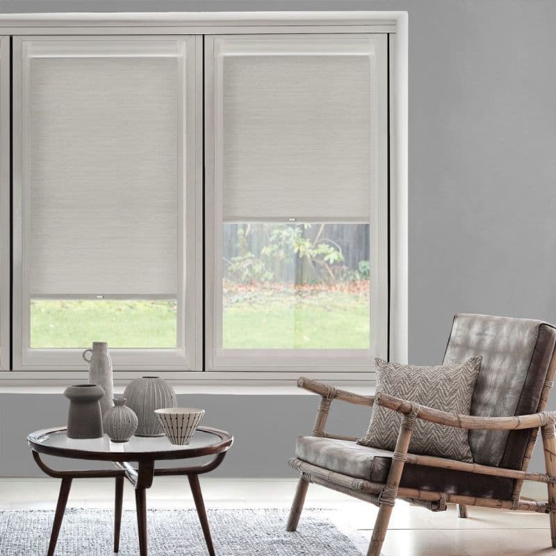 Light beige textured roller blinds, lowered across a two-panel window, partially filtering daylight; living room with gray walls, rattan-framed leather chair, small round table and lawn view.