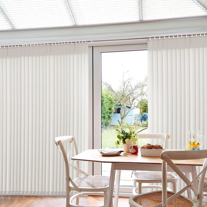 Vertical white fabric vertical blinds cover sliding patio doors, panels mostly closed with a central gap; filtering daylight into a bright dining nook with wooden table, chairs, flowers, garden view.