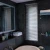 Aluminum horizontal blinds, closed with slats slightly tilted, filtering daylight through a tall narrow window; dark-tiled bathroom surrounding a freestanding tub, mirror, wire shelves holding towels and toiletries.