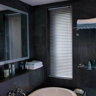 Aluminum horizontal blinds, closed with slats slightly tilted, filtering daylight through a tall narrow window; dark-tiled bathroom surrounding a freestanding tub, mirror, wire shelves holding towels and toiletries.