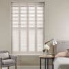 White full-height plantation shutters, four-panel with adjustable louvers, closed and filtering daylight, covering a recessed window; neutral bedroom corner with armchair, bedside table, lamp, and flowers.
