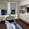 Two gray Roman fabric blinds, lowered with flat folds, partially covering windows and blocking light; modern dining-living room with white table and chairs, orchid, blue sofa, built-in cabinets, dusk.