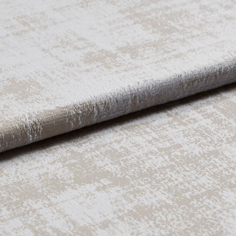 A folded plush velvet-like upholstery fabric with a mottled beige and off-white pattern is layered to reveal pile and edge, shown close-up on a neutral surface under soft studio lighting.