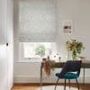 Pale patterned Roman blind lowered over a window, with subtle ikat-diamond fabric; filtering soft daylight onto a desk with mirror, vase of flowers and teal chair.