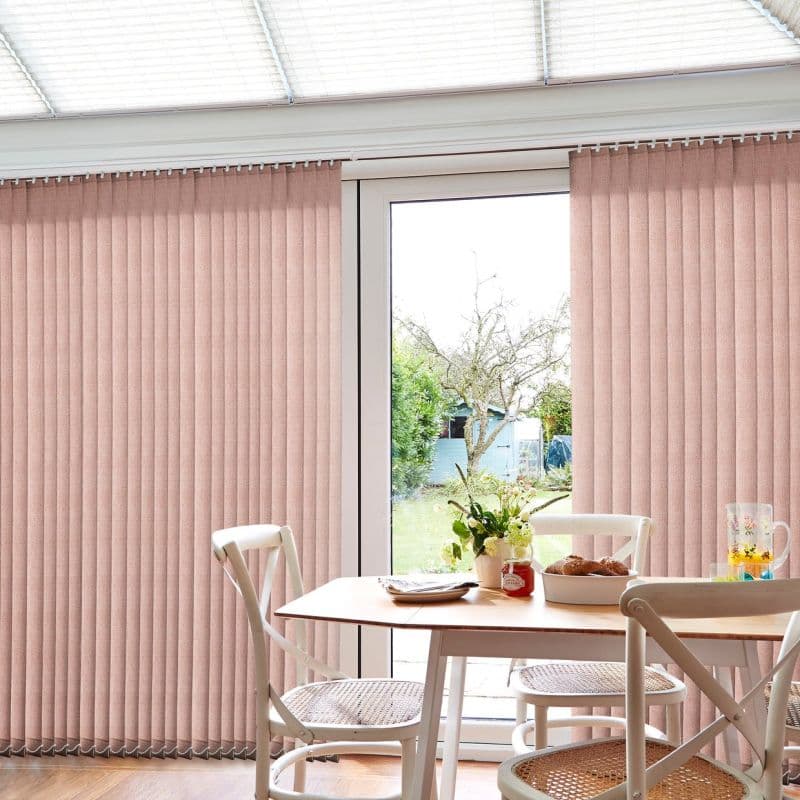 Vertical fabric blinds, blush-pink, spanning ceiling-to-floor and parted at a glass patio door; mostly closed, diffusing daylight. Surrounding dining area with wooden table, cane-seat chairs and garden view beyond.