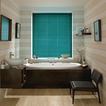Teal horizontal blinds covering a centered window, fully lowered and filtering daylight above a built-in wooden bathtub in a striped-tile bathroom with mirrors, shelves, vase, and a chair.
