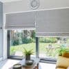 Grey Roman blinds covering sliding glass doors, lowered and overlapping slightly, filtering daylight; a coffee table with potted ferns and a yellow sofa face a sunlit lawn and flowered garden outside.
Text: Greenwich