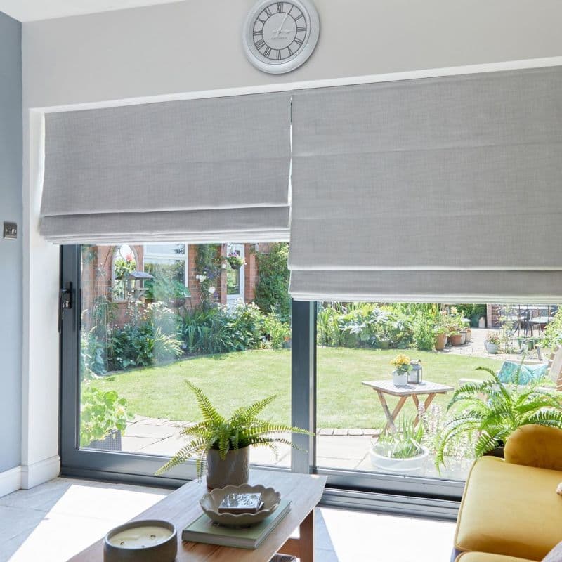 Grey Roman blinds covering sliding glass doors, lowered and overlapping slightly, filtering daylight; a coffee table with potted ferns and a yellow sofa face a sunlit lawn and flowered garden outside.
Text: Greenwich