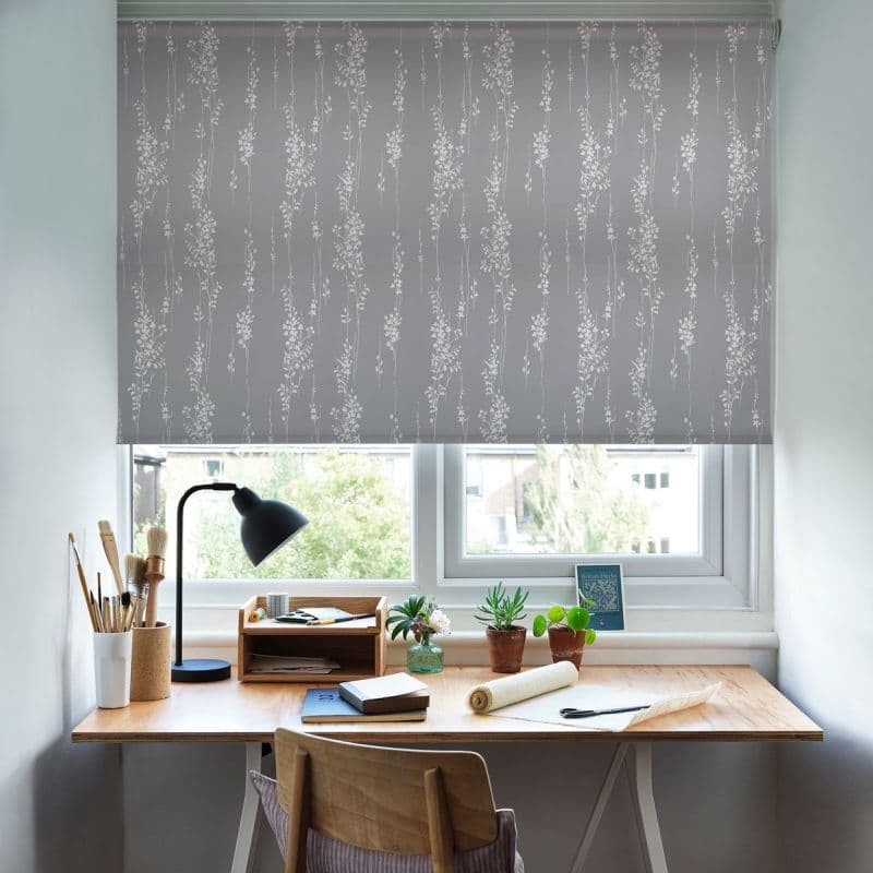 Grey roller blind with white vertical botanical pattern, fully lowered over a double window; daylight-dimmed home workspace with wooden desk, lamp, brushes, notebooks and small potted plants.