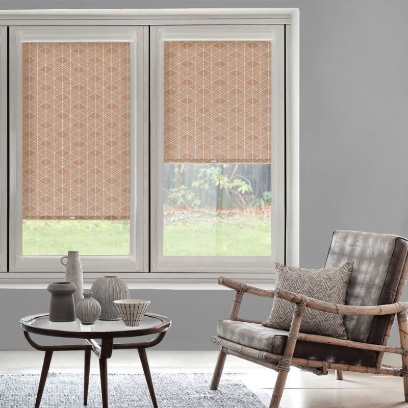 Peach geometric roller blinds across a two-panel window—left fully lowered, right partially lowered—filtering daylight and revealing a lower yard view; gray-walled living room with wicker armchair and ceramic-decorated coffee table.