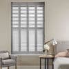White plantation-style shutters covering a tall window, fully closed with slats slightly tilted to admit soft light; bedroom setting with upholstered bed, side table lamp, armchair, neutral walls.