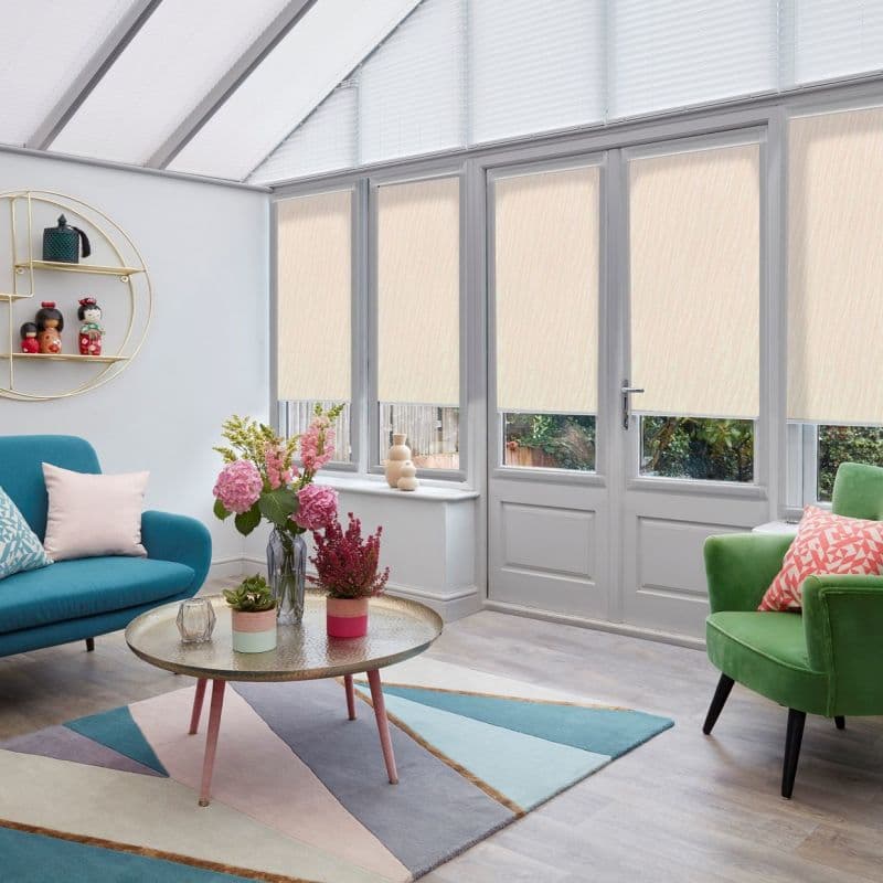 Beige fabric roller blinds spanning glass doors and tall windows, partially lowered and filtering daylight, in a bright conservatory-style living room with teal sofa, green armchair, flowers and geometric rug.