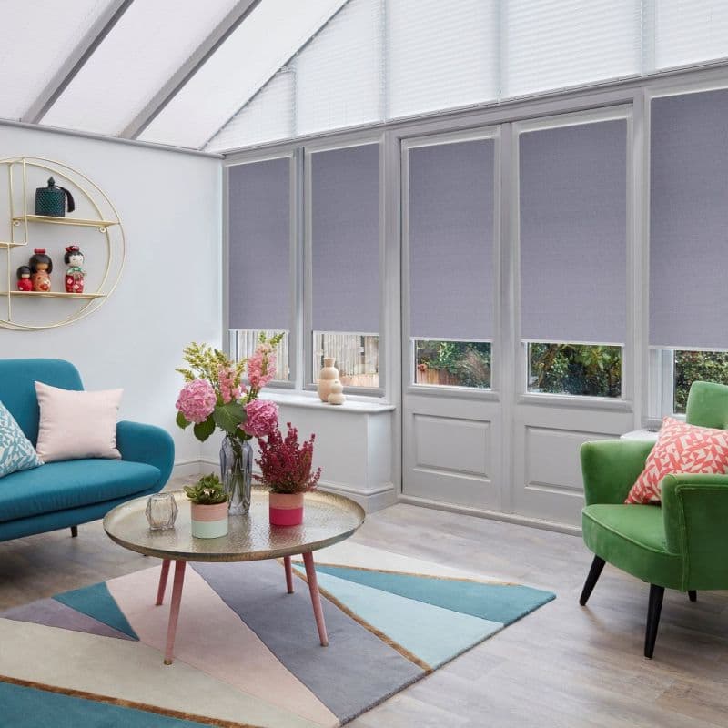 Light grey fabric roller blinds, lowered to cover most of tall rectangular windows and patio door, softly filtering daylight; conservatory-style living room with skylights, teal sofa, green armchair, floral centerpiece.
