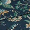 Folded dark teal fabric with botanical print—green leaves and yellow, blue flowers—folded along the center, overlapping to reveal textured weave; close-up on a flat surface under soft studio lighting.