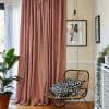 Dusty-rose floor-length curtains with sheer white voile behind, pleated and pooling; filtering daylight from a sliding door — corner living room with rattan chair, black-and-white cushions, plant, hanging bulb, framed 24 7