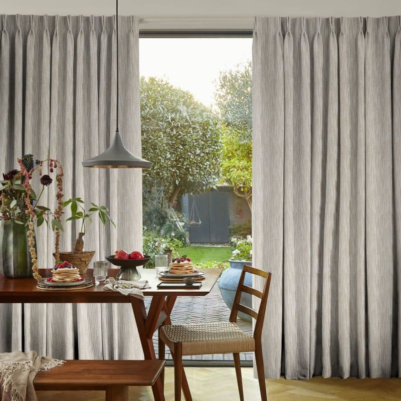 Floor-to-ceiling pleated beige curtains, partially drawn and filtering soft daylight through a central glass door; dining table with plates, pendant lamp and chairs, opening onto a verdant backyard.