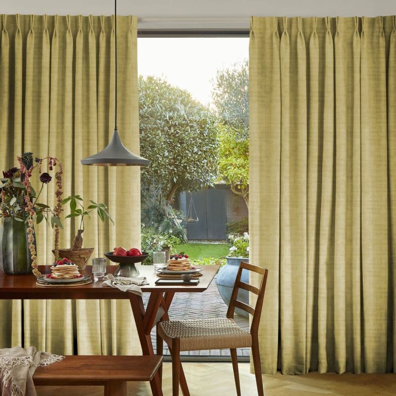 Floor-length pleated beige curtains flank a sliding glass door, partially drawn to reveal a sunlit garden; curtains filter warm light into a dining nook with wooden table, chairs, pendant lamp.