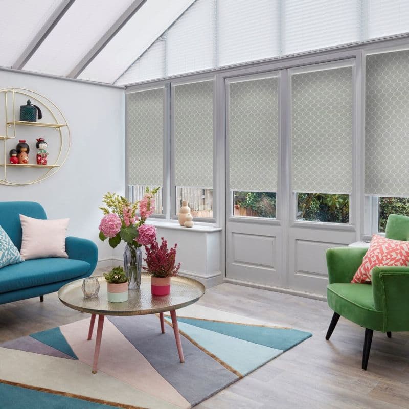 Patterned pale roller blinds partly lowered over tall conservatory windows and doors, filtering daylight; modern sunroom with teal and green armchairs, geometric rug, round coffee table and flower arrangements.