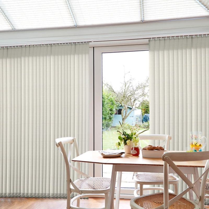 Cream vertical blinds covering a sliding patio door, parted at center to reveal glass; slats mostly closed, filtering daylight; small dining table and chairs in front, garden visible beyond.