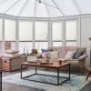 White cellular pleated blinds fitted across tall conservatory windows and roof panels, lowered to filter soft daylight; cozy sunroom with beige sofa, wooden coffee table, pastel throws, and blue rug.