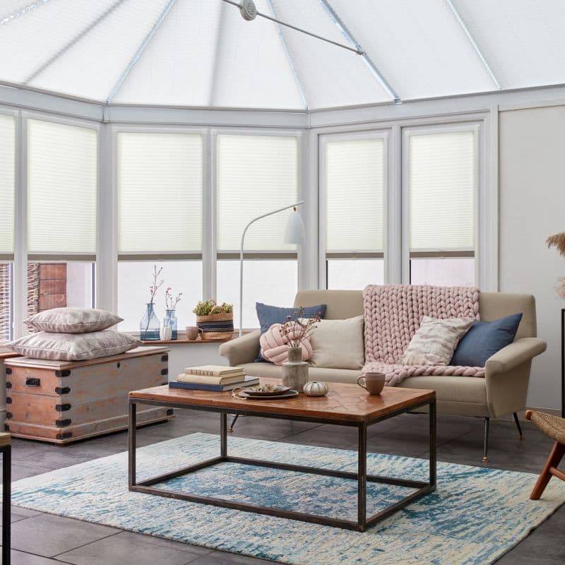 White cellular pleated blinds fitted across tall conservatory windows and roof panels, lowered to filter soft daylight; cozy sunroom with beige sofa, wooden coffee table, pastel throws, and blue rug.