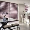 Dual-layer roller blinds with alternating burgundy opaque and sheer horizontal stripes covering floor-to-ceiling windows and a smaller kitchen window; filtering light into striped shadows; modern open-plan kitchen-dining.