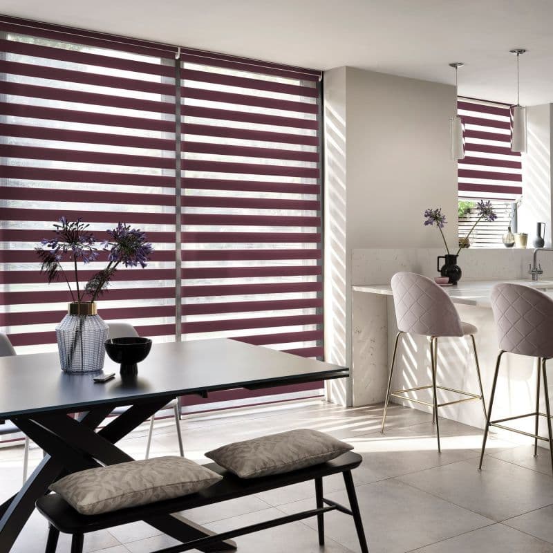 Dual-layer roller blinds with alternating burgundy opaque and sheer horizontal stripes covering floor-to-ceiling windows and a smaller kitchen window; filtering light into striped shadows; modern open-plan kitchen-dining.