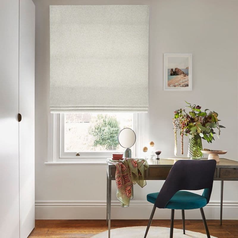 Pale textured Roman blind lowered over a sash window, folded at bottom and filtering daylight; above a wooden desk with chair, mirror and vase with flowers in a neutral room.