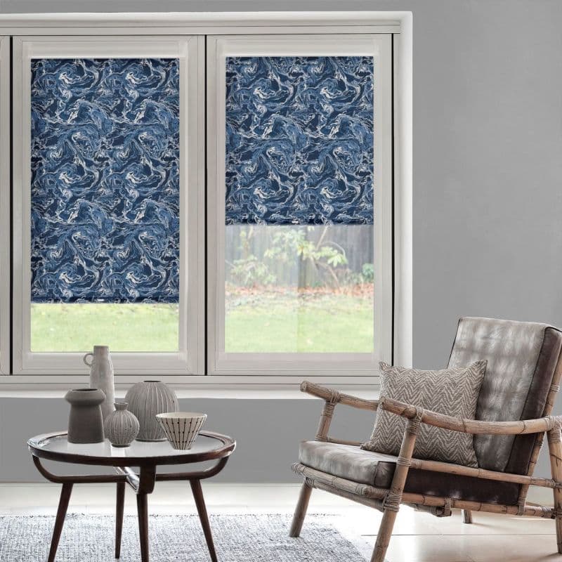 Blue-and-white swirling roller blinds on two tall white-framed windows — left fully lowered, right partially lowered — filtering light; neutral living room with rattan armchair, small round table and lawn view.